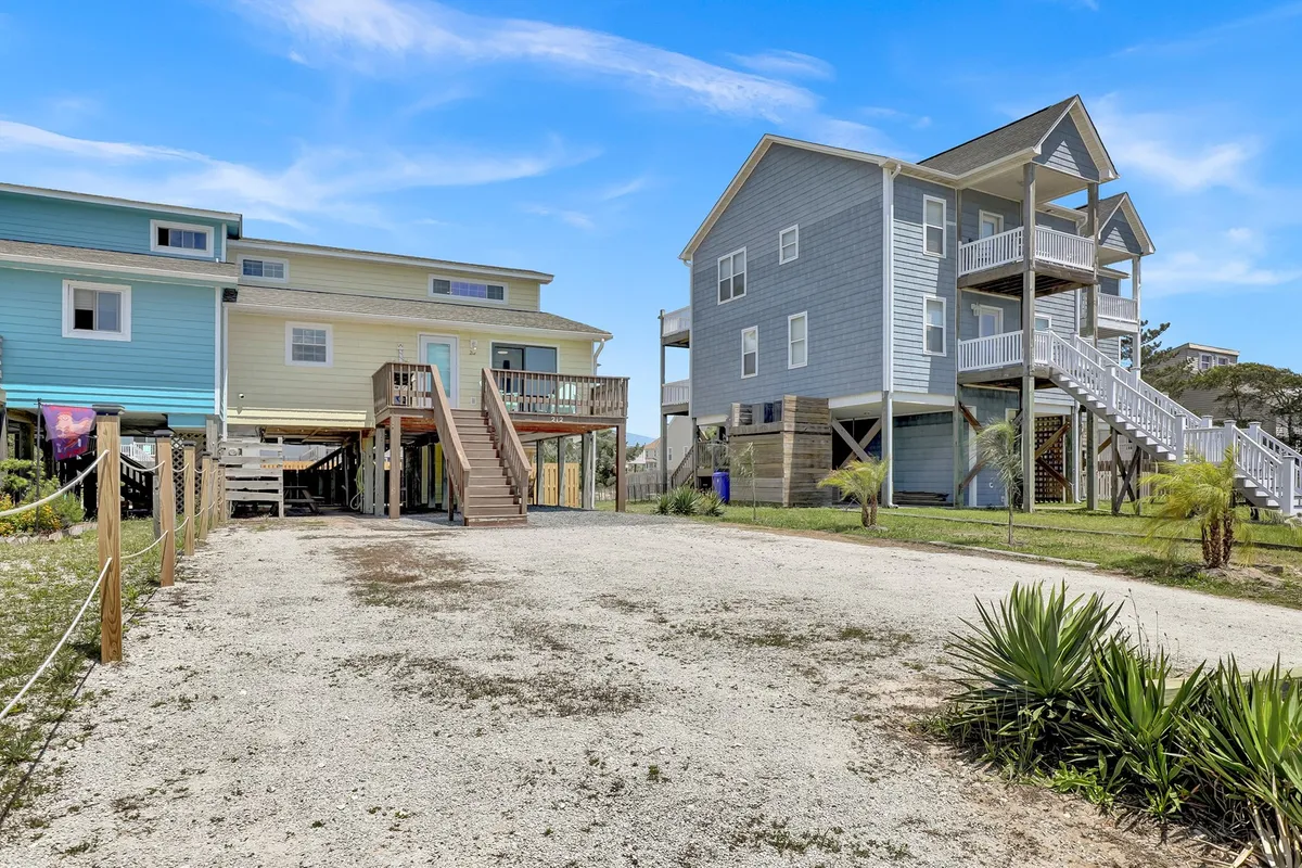 This image features two multi-story beach houses set along a gravel driveway bordered by lush greenery. The homes have spacious decks perfect for enjoying sunset views, while the setting is casual and welcoming, ideal for family gatherings or evening barbecues.