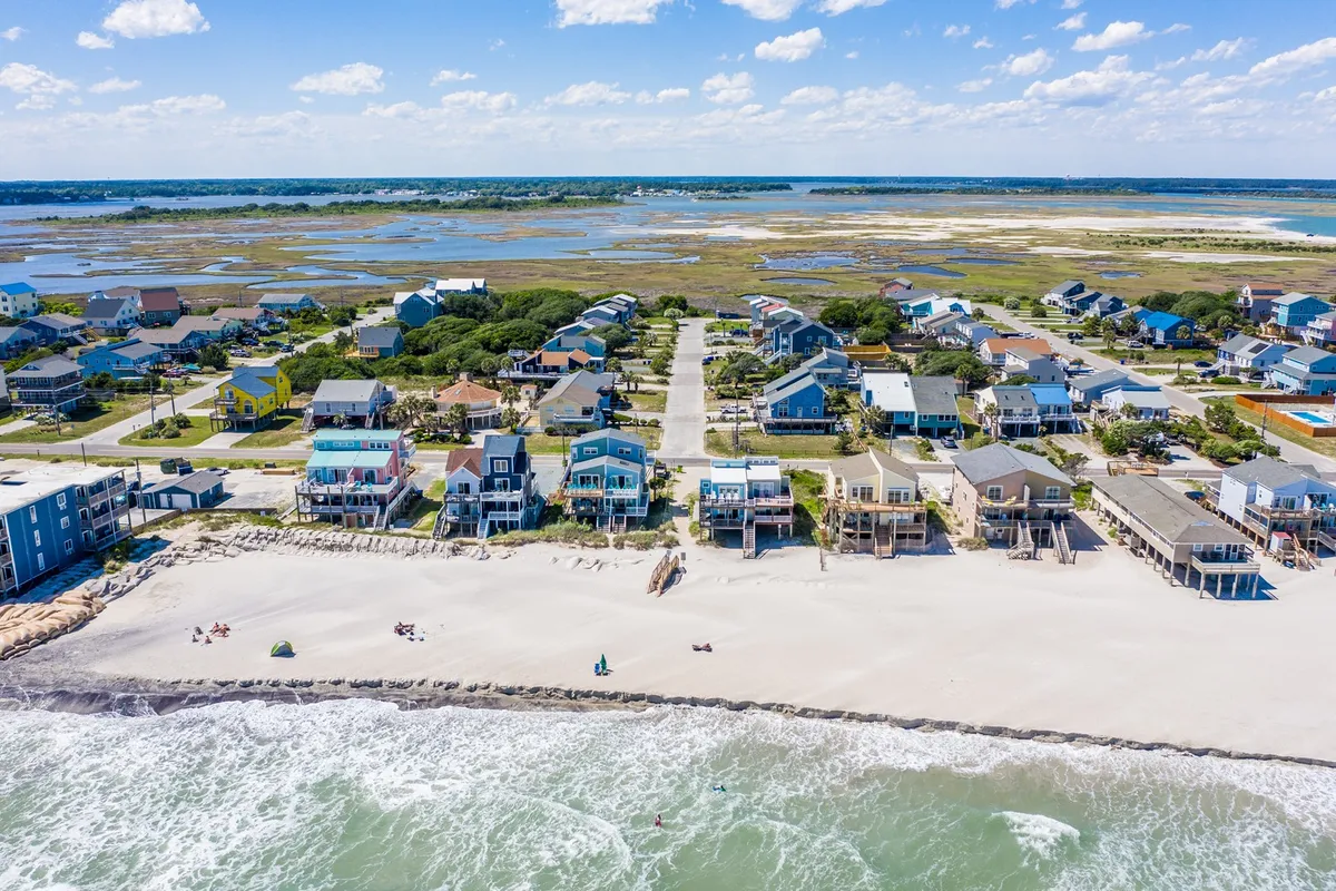 This image features a panoramic view of colorful beachfront houses lining the shore, with the ocean waves gently meeting the sandy beach and a stretch of marshland visible under a broad blue sky. The scene is open and inviting, clearly emphasizing the proximity of the homes to the water and the accessibility of the beach for walks.