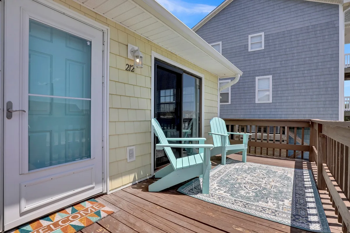 A close-up view of a welcoming entryway featuring blue-green Adirondack chairs, a decorative rug, and a friendly welcome mat. The scene suggests a relaxing spot for morning coffee or unwinding at sunset, framed by thoughtful, homey details.