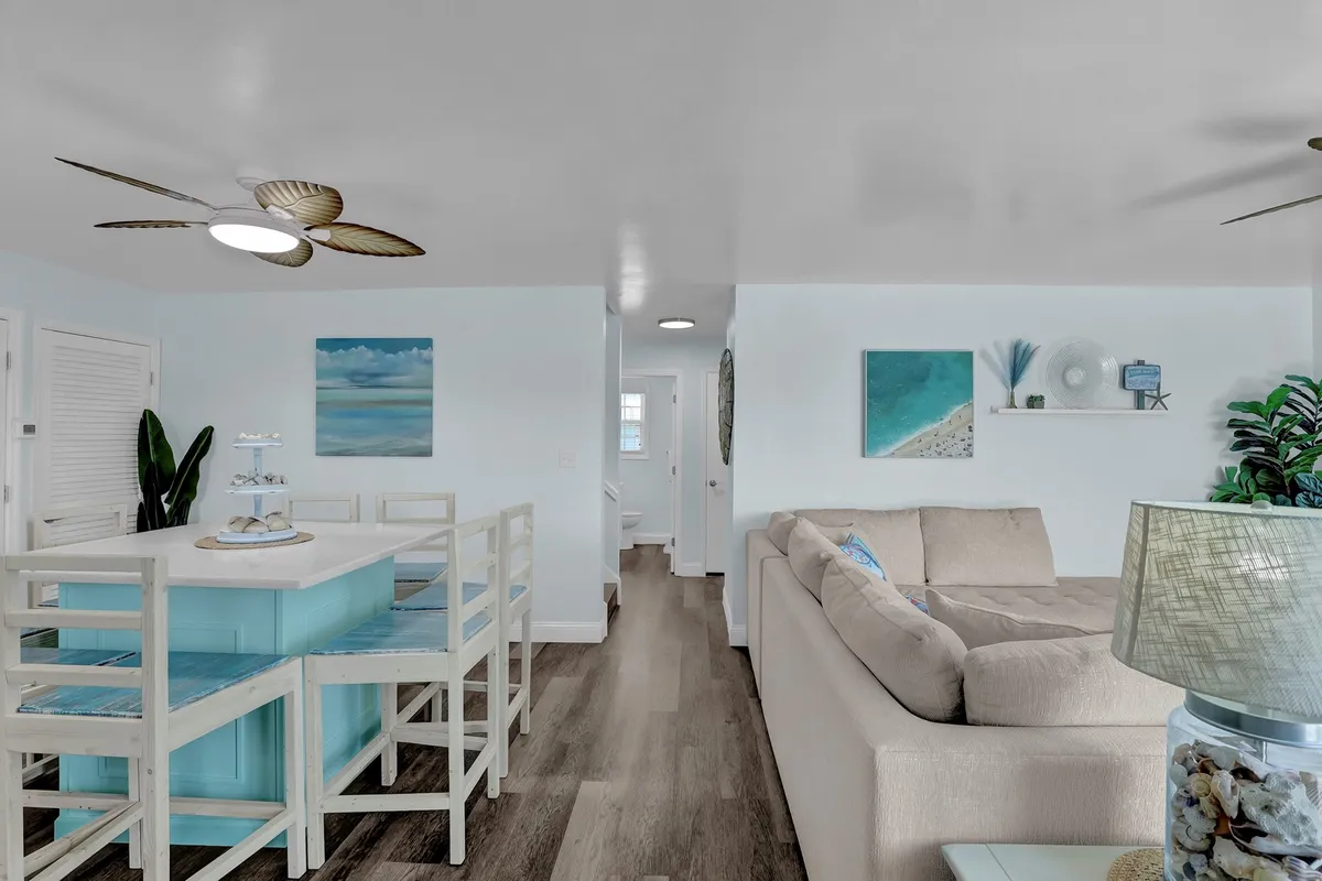 A seamless open-concept space that combines a light blue dining table, a plush beige sofa, and tropical ocean-themed artwork. The atmosphere is casual and inviting, with ample soft seating and subtle coastal touches throughout.