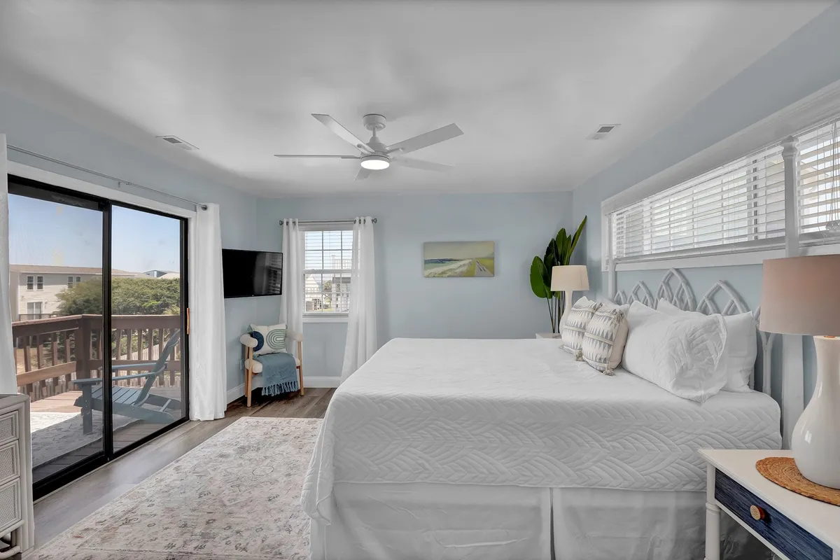 A spacious, sunlit primary bedroom with a large bed dressed in crisp white linens. A sliding glass door opens directly onto a private balcony, allowing sunlight to stream in and offering a glimpse of the outdoors. The space is thoughtfully decorated with a modern lamp and tasteful artwork, emphasizing comfort and style.