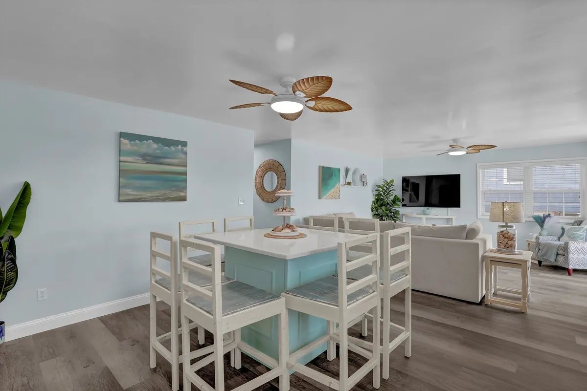A bright dining area featuring a spacious table with white chairs, a ceiling fan with a leaf motif, ocean-themed wall art, a green plant in the corner, and a television mounted on the wall. The setting is cheerful and ready for family meals or group gatherings.