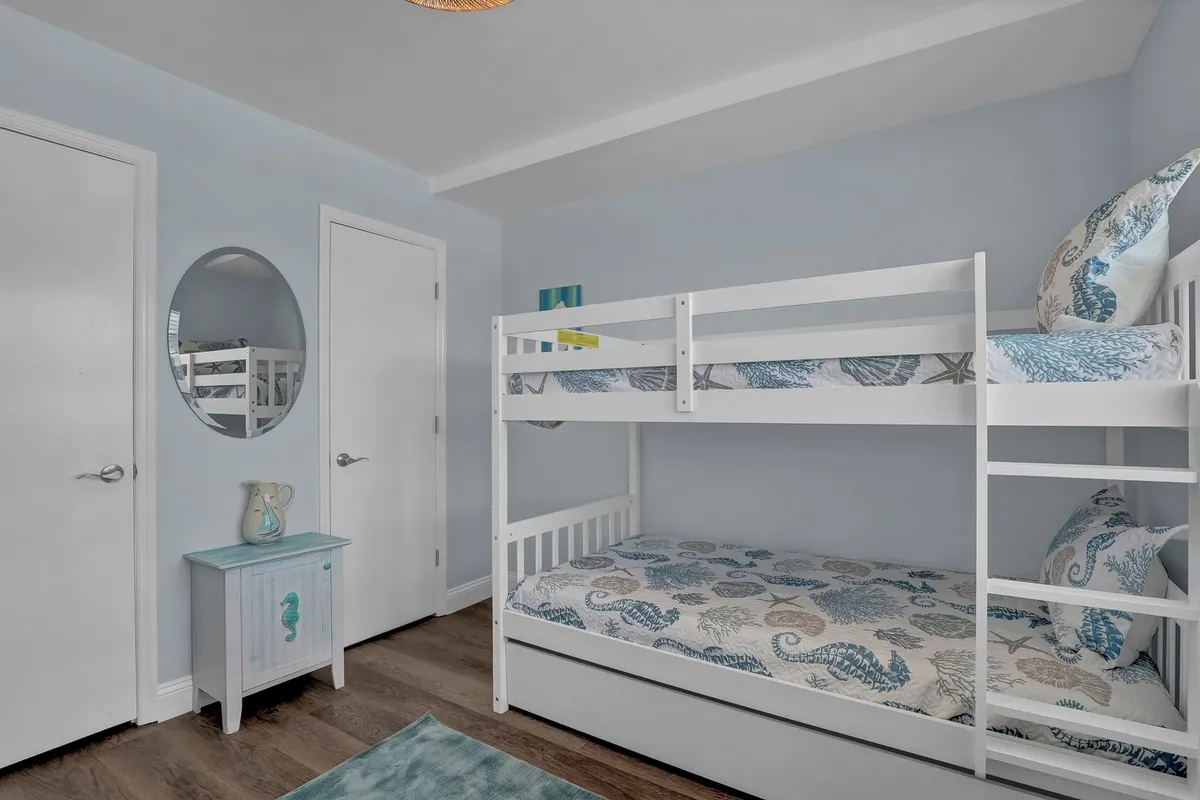 A playful bunk room featuring a set of sturdy, stylish bunk beds adorned with decorative pillows. A cheerful blue nightstand, a mirror, and other playful accents create a cozy and fun environment ideal for kids or extra guests.