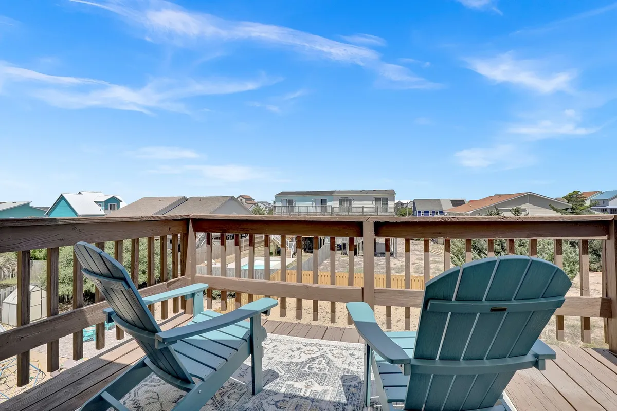 This image features a tranquil deck with adirondack chairs, a deck railing, an outdoor rug, and a view of the neighborhood beyond. The space is designed for relaxing and socializing, offering an inviting transition from indoor to outdoor living.