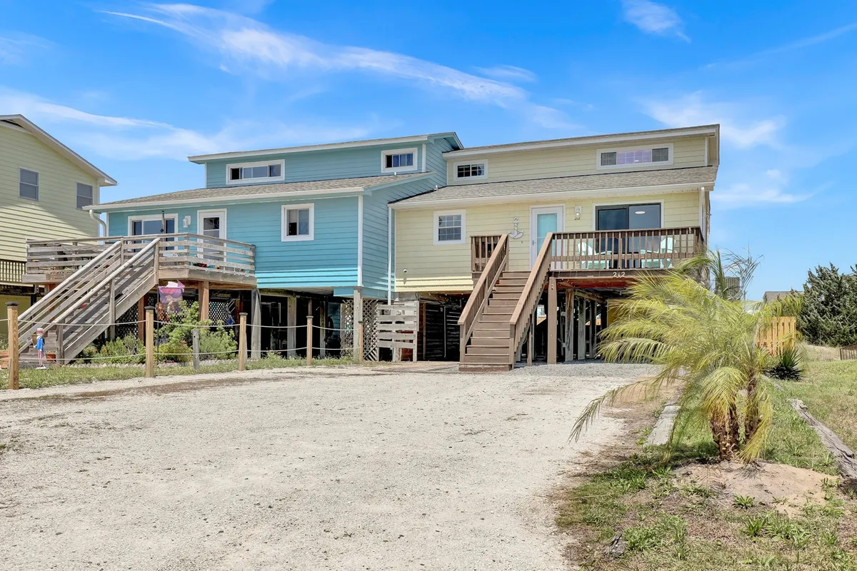 This exterior image showcases a charming row of colorful houses with inviting stairs leading up and lush green landscaping. The scene conveys a lively, welcoming atmosphere, with people gathering for outdoor barbecues, relaxing on porches, and families enjoying quality time together. The sunlight suggests the potential for beautiful sunset views from the deck, connecting to the section's emphasis on gatherings and sunset picnics.