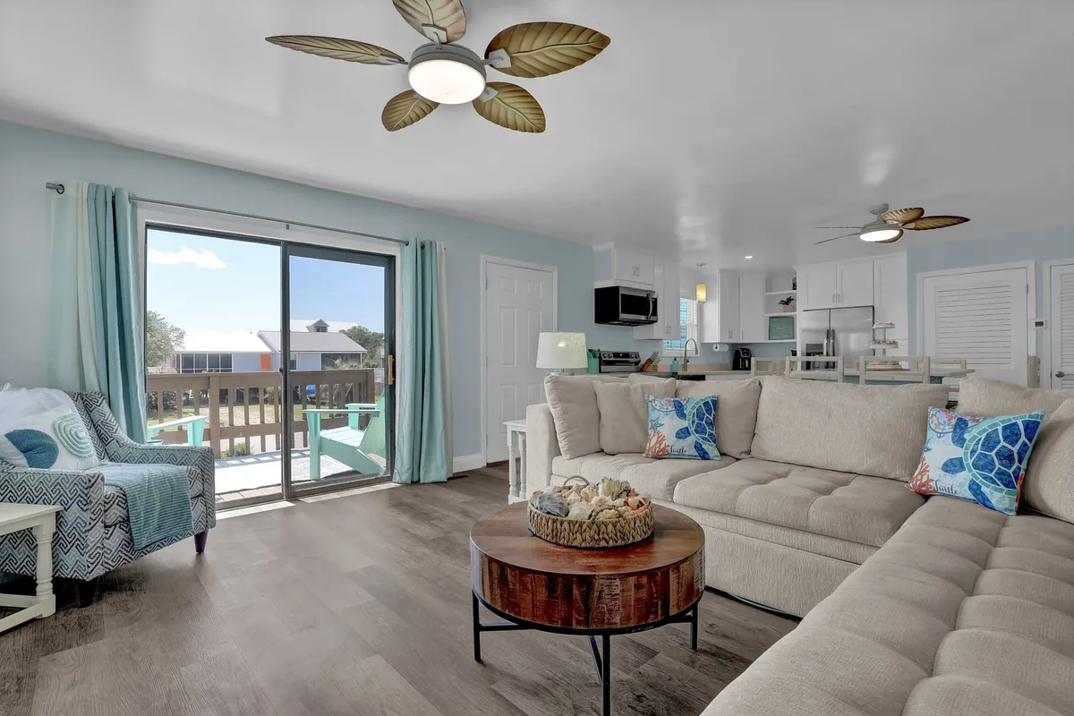 The hero image showcases a spacious and sunlit living area featuring a large sectional sofa, a cozy dining space, and sliding glass doors that open to a deck with sunset views. The inviting setup hints at relaxing family gatherings, movie nights, and effortless indoor-outdoor living.
